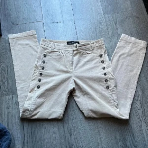 Beiga manchesterbyxor Kathleen Madden stl 38 - Snygga beiga manchesterbyxor från Kathleen Madden med raka ben och dekorativa knapprader längs sidorna och på bakfickorna. Tunt Manchestertyg.