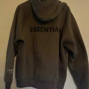 Grå Essentials Fear of God hoodie S - Säljer en grå hoodie från Essentials Fear of God i storlek S men passar också till personer med storlek M. Tröjan har dragkedja, huva och stora fickor framtill. Tillverkad i 100% bomull och har tydlig logga på ryggen. Perfekt för en chill och stilren look. 