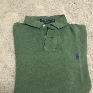 Grön pikétröja från Polo Ralph Lauren - Snygg grön pikétröja från Polo Ralph Lauren med klassisk krage och två knappar framtill. Tröjan har det ikoniska lila Polo-logot broderat på bröstet. Perfekt för dig som gillar stilrena och tidlösa plagg.
