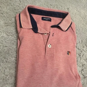 Rosa pikétröja från Jack & Jones - Snygg rosa pikétröja från Jack & Jones med mörkblå krage och detaljer. Tröjan har klassisk pikéknäppning med två knappar och en liten broderad logga på bröstet. Perfekt för en stilren och avslappnad look.