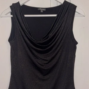 Glittrig svart ärmlös topp Street One - Svart glittrig topp från Street One med snygg draperad halsringning. Toppen är ärmlös och har en lätt skimrande finish som ger en cool look. Kan bäras både med cowlneck eller offshoulder-stil. Perfekt för dig som gillar att sticka ut lite extra.