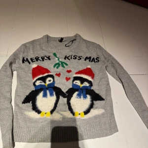 Grå jultröja med pingviner och text - Grå stickad jultröja med motiv av två söta pingviner i tomteluva och blå halsduk, röda hjärtan och texten 'Merry Kiss-mas' framtill. Tröjan har rund halsringning och långa ärmar. Perfekt för julens alla tillfällen!