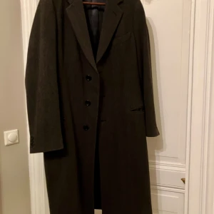 Armani Collezioni lång rock i ull - Säljer en lång, klassisk höst-/vinterrock från Armani Collezioni i mörkgrått fiskbensmönster. Rocken har tre knappar framtill, spetsiga slag och fickor med lock. Tillverkad i ull med slät fodring. Perfekt för dig som är lite smalare om axlarna då den sitter perfekt. 