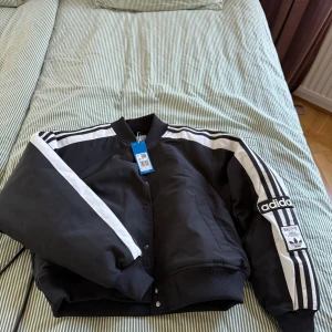 Svart bomberjacka från Adidas - Snygg svart bomberjacka från Adidas, jackan är oanvänd med prislapp kvar. Inköpt för 1.400, säljer för 500. Den är stor i storleken 
