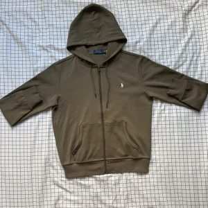 Olivgrön hoodie från Polo Ralph Lauren - Olivgrön hoodie från Polo Ralph Lauren med dragkedja, huva och fickor framtill. Klassisk broderad logga på bröstet och trekvartsärmar. Tillverkad i mjuk bomull, perfekt för chill dagar eller när du vill ha en clean look.