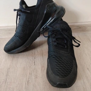 Svarta Nike Air Max 270 sneakers - Säljer ett par helsvarta Nike Air Max 270 sneakers med snygg mesh-överdel och tydlig Air-dämpning i hälen. Skorna har snörning och en sportig vibe, perfekta för dig som gillar streetstyle. Modellen är lätt och andas bra, med diskreta Nike-loggor på plösen och hälen.