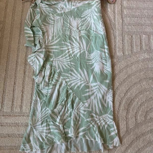 Grön omlottkjol med volang - Snygg långkjol i ljusgrönt och vitt med tropiskt bladmönster. Kjolen har omlottmodell och volangdetaljer längs kanten, vilket ger en flowig och somrig känsla. Perfekt för varma dagar och enkel att styla med en enkel topp.