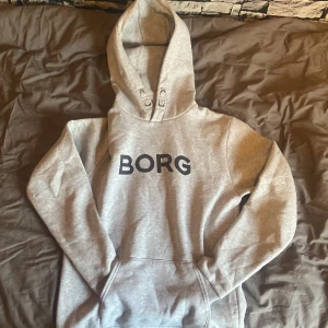 Grå hoodie från Björn Borg - Snygg grå hoodie från Björn Borg med svart BORG-tryck på bröstet. Klassisk modell med huva och känguruficka framtill. Perfekt för chill dagar eller när du vill ha en enkel och stilren look.