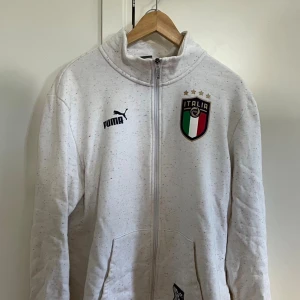 Vit Italia ziptröja från Puma - Säljer en vit ziptröja från Puma med Italiens landslagsemblem på bröstet och stor ITALIA-tryck på ryggen. Tröjan har hög krage, hel dragkedja och två fickor framtill. Materialet är mjukt och bekvämt, perfekt för fotbollsfans.