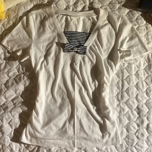 Vit t-shirt med randig rosett - Vit t-shirt med v-ringning och korta ärmar. Framtill finns en blåvit randig detalj och en söt rosett vid halsen. Gjord i mjuk bomull, perfekt för en chill och stilren look.