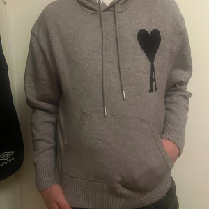 Ami Paris  - Snygg grå hoodie från Ami Alexandre Mattiussi med svart hjärta och A på bröstet. Tröjan har huva med snörning, ribbade muddar och en stor magficka. Perfekt för dig som gillar stilrena streetwear-plagg med coola detaljer.