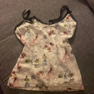 Mönstrad linnetopp med spetsdetaljer - Supersöt linnetopp med vintageinspirerat mönster av kvinnor i klänningar och blommor. Toppen är vit med inslag av rött, blått, grönt och svart, och har tunna axelband med svarta spetsdetaljer. Materialet är glittrigt och har en lätt spetsstruktur för extra wow-känsla.