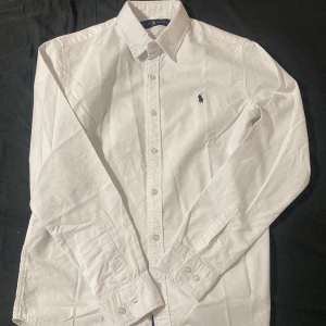 Vit skjorta från Ralph Lauren S - Klassisk vit skjorta från Ralph Lauren med blå broderad logga på bröstet. Skjortan har lång ärm, knappar hela vägen och krage. Tillverkad i bomull för en skön och stilren känsla. Perfekt för dig som gillar en clean och tidlös look.