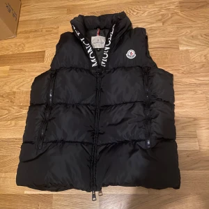 Svart dunväst från Moncler - Svart dunväst från Moncler med hög krage och vit broderad logga längs dragkedjan. Västen har två fickor med dragkedja och klassisk Moncler-patch på bröstet. Perfekt för lager-på-lager och streetstyle.