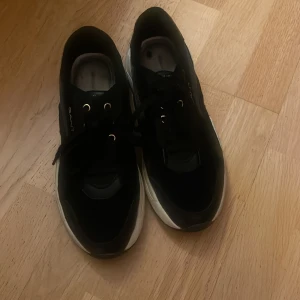 Svarta sneakers från Gant - Snygga svarta sneakers från Gant med vita, chunky sulor och gulddetaljer vid snörningen. Skorna har en sportig vibe med rund tå och Gant-logga på sidan och hälen. Ovandelen är i en mix av skinn och syntetmaterial. Perfekta för dig som gillar stilrena och bekväma sneakers.