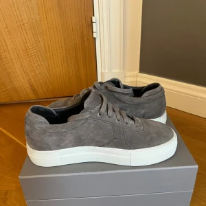 Grå mockasneakers från Axel Arigato - Snygga grå sneakers från Axel Arigato i mjuk mocka med vit platt sula. Skorna har klassisk rund tå, snörning och stilren design som passar till allt. Perfekta för dig som gillar minimalistisk och clean stil.