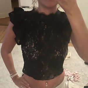 Svart spetsig croppad blus med volang - Säljer en svart croppad blus i spets med volangdetaljer på axlarna och hög hals. Toppen är ärmlös och har en transparent look som ger en edgy vibe. Perfekt för dig som vill sticka ut med en snygg och trendig topp.