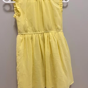 Gul volangklänning från Name It - Söt gul klänning från Name It i storlek 128. Klänningen har volangdetaljer vid axlarna och är ärmlös med en luftig, lätt kjol. Perfekt för varma dagar och har en elastisk midja för skön passform.