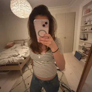 Randig topp från Holly & Whyte - Säljer en vit och marinblå randig topp från Holly & Whyte by Lindex. Modellen är croppad med korta ärmar, rundad ringning och tre dekorativa knappar framtill. Toppens material är ribbad bomull som ger en skön och stretchig känsla.