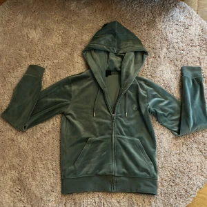 Grön velour hoodie från Juicy Couture - Mjuk grön hoodie i velour från Juicy Couture med dragkedja framtill, huva med snörning och två fickor. Klassisk logga broderad på bröstet. Perfekt för chill dagar och snygg att matcha med jeans eller mjukisbyxor.