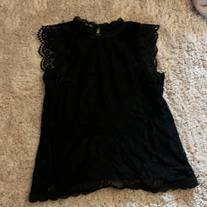 Svart spetsblus med volangärm - Supersnygg svart blus i spets med ärmlös design och volangdetaljer vid axlarna. Hög hals med liten spetskant och knäppning bak i nacken. Perfekt för dig som vill ha en feminin och trendig look.