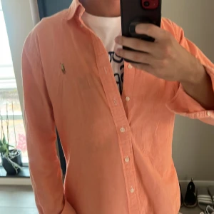 Persikofärgad skjorta från Ralph Lauren - Säljer en persikofärgad skjorta från Ralph Lauren med klassisk krage och knäppning framtill. Skjortan har lång ärm och den ikoniska broderade loggan på bröstet. Tillverkad i mjuk bomull som känns skön mot huden. Perfekt för dig som vill ha en chill och stilren look. Mitt pris 349kr