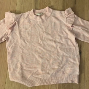 Ljusrosa stickad tröja med volang - Supersöt ljusrosa stickad tröja från Uniq Studio med volangdetaljer på axlarna och ribbade muddar. Perfekt för dig som gillar en feminin och trendig look. Långärmad och mjuk, passar perfekt till jeans eller kjol.