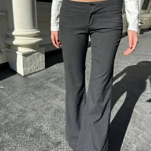 Grå randiga kostymbyxor bootcut - Snygga grå kostymbyxor med tunna vita ränder och bootcut-snitt. Byxorna har låg midja och klassisk passform, perfekta för en trendig och stilren look. Materialet känns dressat och passar till många olika outfits.