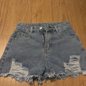Högmidjade slitna jeansshorts - Säljer ett par ljusblå högmidjade jeansshorts med råa, fransiga kanter och slitningar framtill. Klassisk femficksmodell med dragkedja och knapp. Perfekta för sommaren och ger en avslappnad vibe.