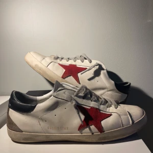 Golden Goose skor - Snygga vita sneakers från Golden Goose med ikonisk röd stjärna på sidan, grå snörning och svart hälparti. Skorna är i skinn med beige mockadetaljer vid hälen och har en platt sula i gummi. Perfekta för dig som gillar streetstyle och vill sticka ut. Backsidan är inte svart den är grön ljuset på första bilden gör den mörkgröna svart 