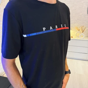Svart t-shirt med Paris-tryck - Svart t-shirt med tryck där det står 'PARIS' och en linje i blått, vitt och rött över bröstet. Klassisk passform med rund hals och korta ärmar. Perfekt för dig som gillar enkel men snygg streetstil. Passar bra som oversize modell.