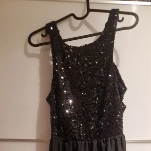 Svart långklänning med paljetter - Svart långklänning med glittriga paljetter på axelband och överdel. Klänningen har öppen rygg med dekorativa band och ett skirt, flödande tyg i kjolen. Perfekt för dig som vill sticka ut med en glamorös look. Materialet känns lätt och elegant.
