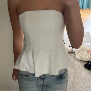 Vit topp zara  - Säljer en snygg vit korsett-topp med axelbandslös design och utsvängd nederkant. Toppen har en figurnära passform och markerade sömmar som ger en stilren siluett. Perfekt att matcha med jeans eller kjol för en trendig look.