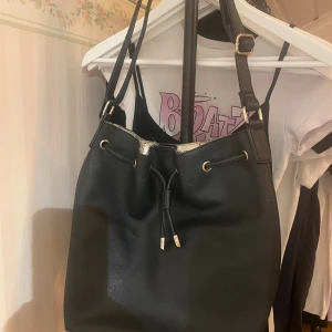 Svart väska med gulddetaljer - Snygg svart väska med justerbar axelrem och guldfärgade metalldetaljer. Väskan har snörning upptill och insida i metallicguld. Perfekt för dig som vill ha en stilren och rymlig väska med coola detaljer. Jag fick den av min kusin förut och den har gått sönder lite upptill vid kanten men annars är den hel.