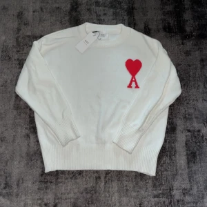 Vit stickad tröja från Ami Paris - Vit stickad tröja från Ami Paris med röd hjärtlogga och bokstaven A broderad på bröstet. Tröjan har rund hals, ribbade muddar och är långärmad. Perfekt för dig som gillar stilrena plagg med en twist.