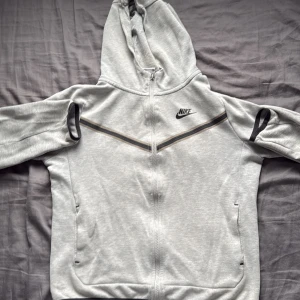 Grå hoodie från Nike med dragkedja - Säljer en ljusgrå hoodie från Nike med hel dragkedja och huva. Tröjan har svarta detaljer, Nike-logga på bröstet och två fickor framtill. Perfekt för chill eller träning. Mjuk och skön bomullsblandning med långa ärmar. Den har några skador som du kan se i bilderna 
