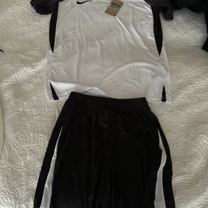Nike Dri-Fit svartvit fotbollströja med shorts  - Snygg svartvit fotbollströja från Nike med Dri-Fit-teknologi. Tröjan har korta ärmar och är tillverkad i ett lätt och ventilerande material som passar perfekt för intensiva matcher. Klassisk design med svart Nike-logga på bröstet.