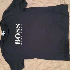Svart t-shirt från Hugo Boss - Svart t-shirt från Hugo Boss med stort vitt BOSS-tryck på bröstet. Klassisk passform med rund hals och korta ärmar. Tillverkad i mjuk bomull som känns skön mot huden. Perfekt för dig som gillar stilrena och snygga logotishirts. Den har ett litet hål på vänster axel men det syns knappt och går att sy, men det behövs egentligen inte