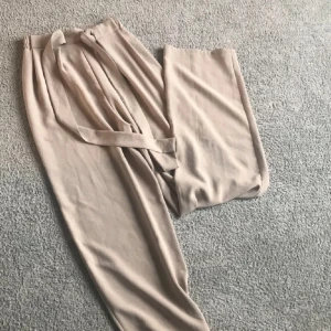 Beiga vida byxor med knytband - Snygga beiga byxor med breda ben och elastisk midja. Byxorna har ett matchande knytband i midjan för extra stilpoäng. Tillverkade i ett mjukt och lätt material som känns skönt mot huden. Perfekta för en chill och avslappnad look.