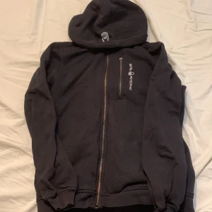 Svart hoodie från Sail Racing med dragkedja - Svart hoodie från Sail Racing med hel dragkedja framtill och broderad logga på bröstet. Tröjan har huva, lång ärm och en bröstficka med dragkedja. Mjuk insida och klassisk passform, perfekt för kyliga dagar.