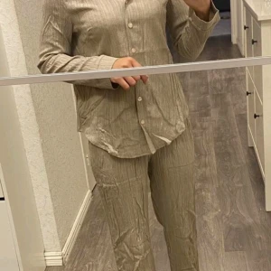 Beige skjorta i krinklad bomull - Säljer ett beige set i krinklad bomull med lång ärm och knappar framtill. Skjortan har en avslappnad passform och klassisk krage. Perfekt för dig som gillar en chill och stilren look.