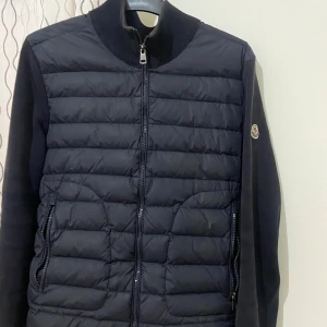 Cardigan Moncler - Snygg  mörklblå cardigan från Moncler i storlek S. Jackan har quiltad framsida med dragkedja, ribbad krage och ärmar i stickad kvalitet. Klassisk Moncler-logga på ärmen och två dragkedjeförsedda fickor framtill. Perfekt för kyliga dagar.