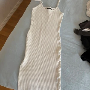 Vit ribbad ärmlös långklänning - Säljer en vit ribbad långklänning med ärmlös design och rund hals. Klänningen har en tight passform och är tillverkad i ett stretchigt material som sitter snyggt på kroppen. Perfekt för dig som gillar en clean och stilren look.