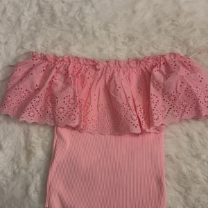 Rosa offshoulder topp med volang - Supersöt rosa offshoulder topp med bred volang upptill och broderade blommor. Toppen är ribbad nedtill och har en axelbandslös design som ger en somrig vibe. Perfekt för dig som gillar romantiska detaljer och en feminin look. Tröjan är köpt utomlands!