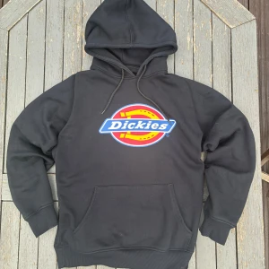 Svart Dickies hoodie med logga - Svart hoodie från Dickies i storlek small med stor färgglad logga på bröstet. Tröjan har huva med snörning och en klassisk känguruficka framtill. Mjuk insida och ribbade muddar vid ärmslut och nederkant. Perfekt för en chill och avslappnad stil.