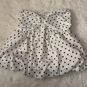 Vit prickig bandeau tubtopp - Supersöt vit bandeau tubtopp med svarta prickar och twistad detalj framtill. Toppen har en luftig, voluminös passform och smockad rygg för extra komfort. Perfekt för dig som vill sticka ut med en retroinspirerad look.