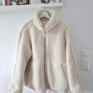 Benvit teddyjacka från H&M, storlek L - Mysig och oversized teddyjacka från H&M i beige med hög krage och dragkedja framtill. Jackan har två sidofickor och är tillverkad i fluffigt teddy-material som håller dig varm under kyliga dagar. Perfekt för dig som gillar en chill och trendig look.
