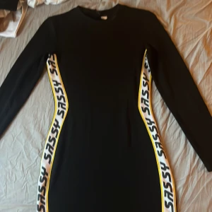 Svart långärmad klänning med SASSY-band - Svart långärmad klänning från H&M med coola vita och gula sidor där det står SASSY i svart text. Klänningen har en tight passform och är perfekt för dig som gillar att sticka ut. Materialet känns mjukt och stretchigt, vilket gör den bekväm att bära hela dagen och kvällen.