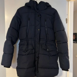 Mörkblå pufferjacka från Everest - Säljer en mörkblå vinterjacka från Everest med huva, dragkedja och knappar framtill. Jackan har två stora fickor med knäppe och två bröstfickor med dragkedja. Perfekt för kalla dagar och har en schysst, lite längre passform. I storlek 164 alltså från barnavdelningen