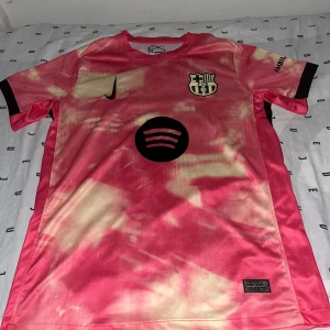 FC Barcelona rosa matchtröja Nike S - FC Barcelona fotbollströja från Nike i rosa och ljusbeige tie-dye stil. Tröjan har klubbmärke, svart Nike-logga och Spotify-logga på bröstet. Tillverkad i lätt Dri-FIT material som andas. Kortärmad modell med rund hals.